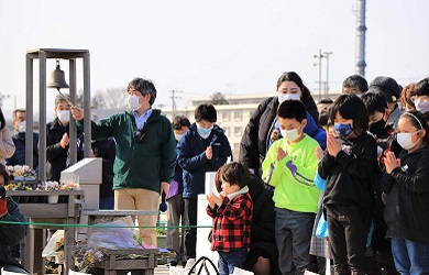 A man, surrounded by a solemn crowd outdoors, rings a bell at a commemorative structure